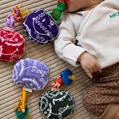 baby met speen in de mond met daaraan een speendoekje met tekst. daarnaast liggen verschillende spenen met speendoekjes. 