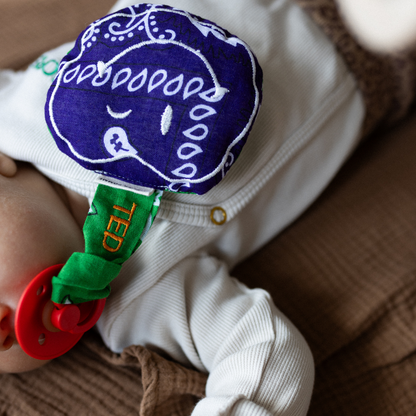 baby ligt met speen in de mond met daaraan een speendoekje met tekst. 
